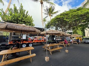 Un camion-restaurant avec un menu et un grill, des tables en bois et des parasols en chaume pour les clients qui souhaitent manger à l’extérieur, et des palmiers en arrière-plan.