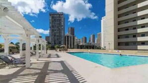 A pool area with a clear sky, a tall building in the background, and a white pergola structure. - King Bed/Renovated/Canal& Mountain View/Walk Beach (Honolulu)