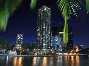 Un immeuble de grande hauteur, comportant de nombreux étages, probablement un hôtel, avec des lumières à chaque étage, situé au bord d’un plan d’eau dont les lumières se reflètent.