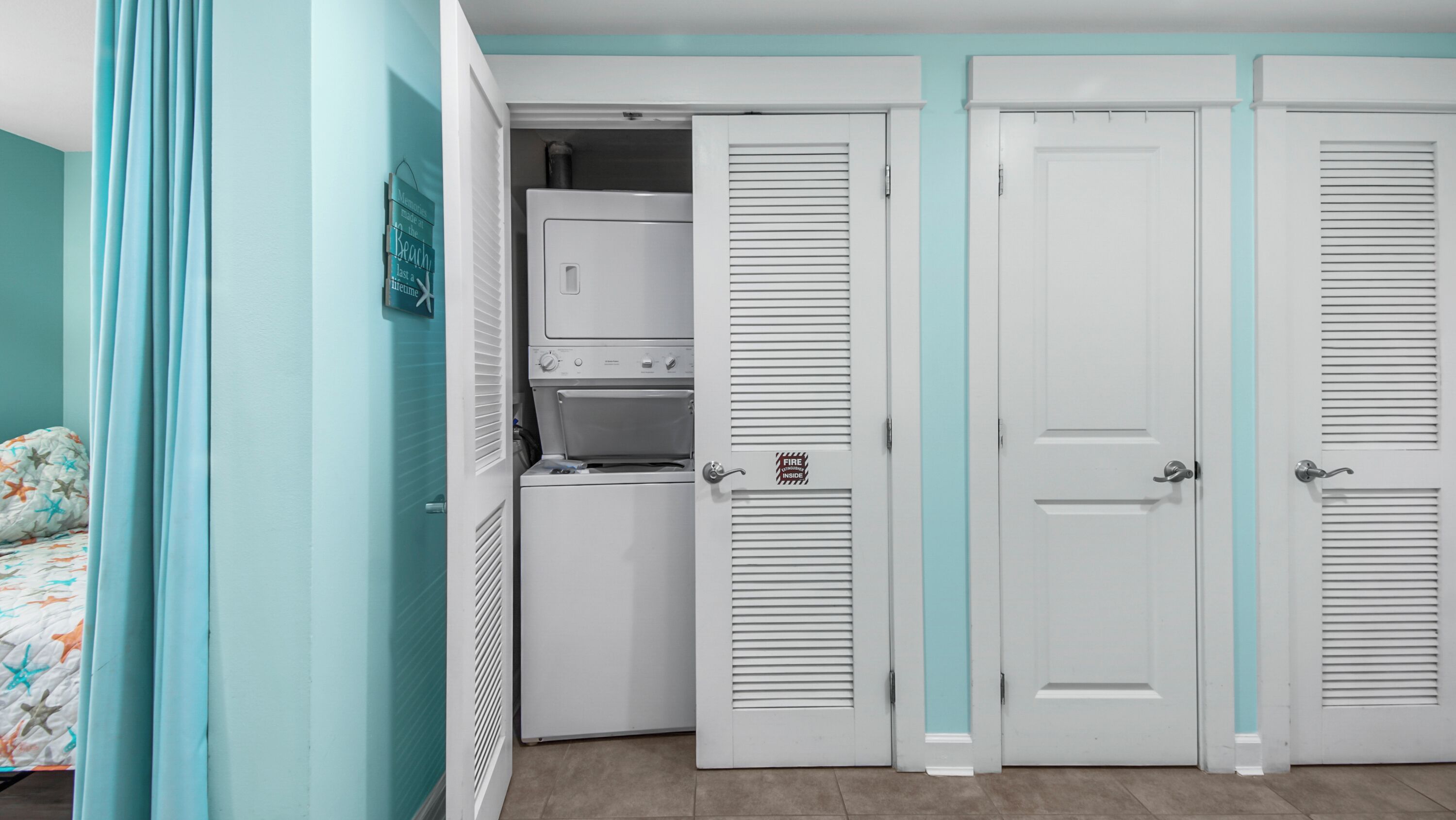 A bedroom with teal walls, a bed with a star-patterned blanket, a washing machine, and a set of white doors with a teal stripe.