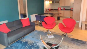 Una sala de estar moderna con un sofá gris, sillas rojas y una mesa de centro. Se ve una cocina al fondo.