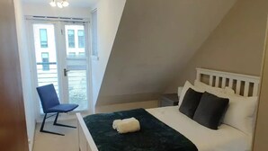Un dormitorio con cama blanca, manta verde oscuro y dos almohadas. Hay una silla y una ventana con persianas.