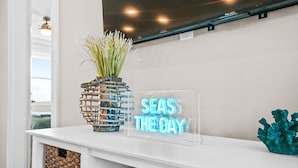 Una consola blanca con un letrero de neón que dice "SEAS THE DAY", un florero con plantas verdes y una escultura de coral azul aguamarina.