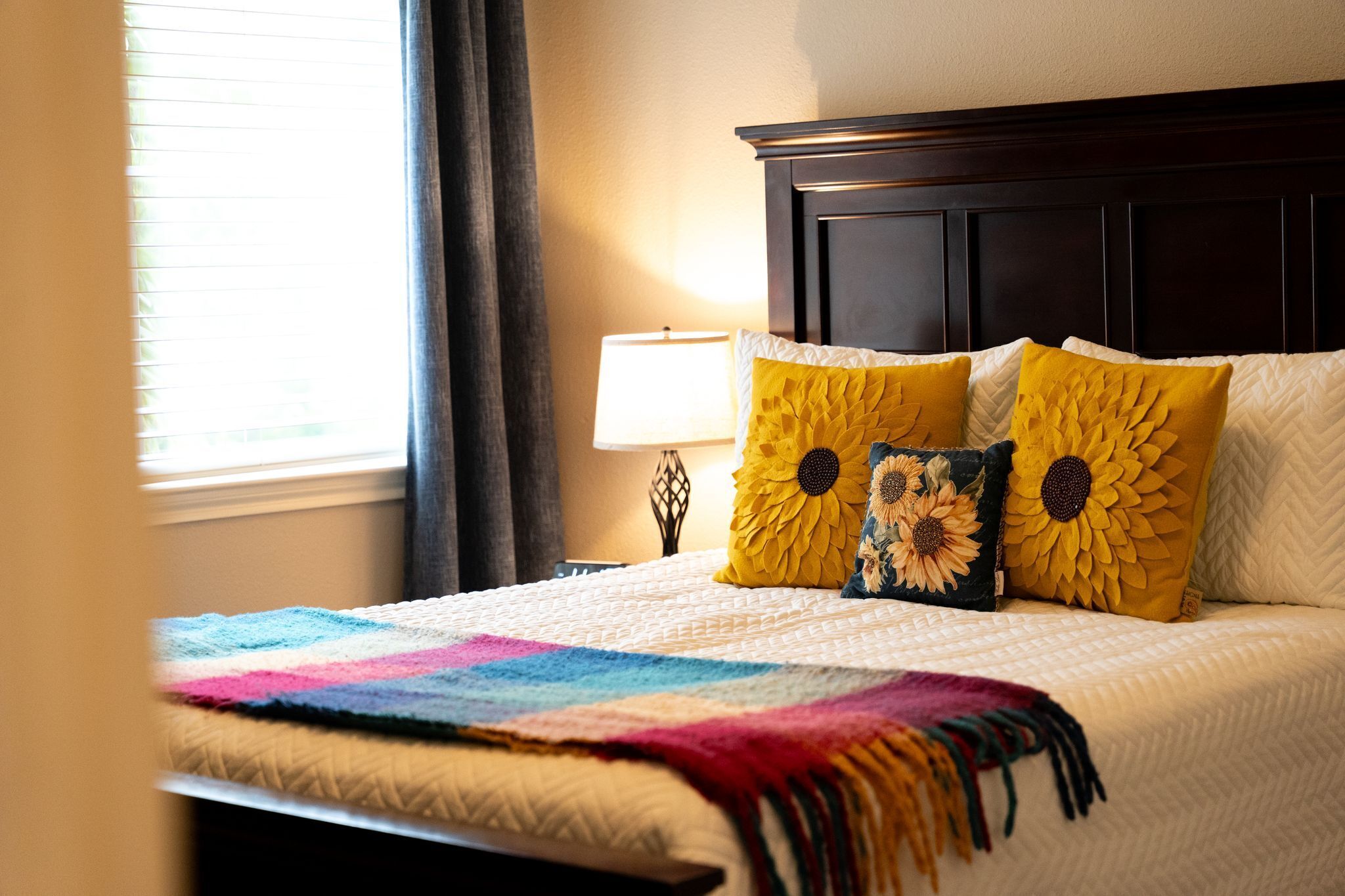 Un lit bien fait, avec une couverture colorée et des oreillers à motifs de tournesols, une tête de lit sombre et une lampe de chevet.