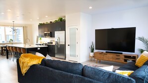 Una sala moderna con un sofá azul oscuro, un televisor plano grande y una cocina con armarios oscuros y electrodomésticos de acero inoxidable.