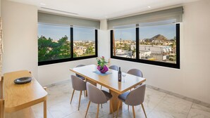 A modern dining area with a wooden table and chairs, large windows with city views, and a small side table with a decorative bowl. - Living in the Heart of Cabos (Cabo San Lucas)