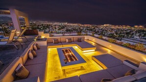 A rooftop terrace with a rectangular fire pit, cushioned seating, and a view of a cityscape at night. - Living in the Heart of Cabos (Cabo San Lucas)