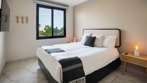 A modern bedroom with a large bed, a nightstand, a bedside lamp, and a window with a view of trees. - Living in the Heart of Cabos (Cabo San Lucas)