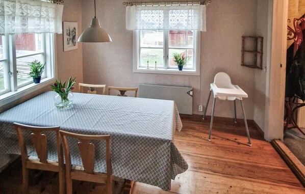 Un comedor con una mesa dispuesta para cuatro, sillas de madera, una lámpara colgante, cortinas de encaje y un ventanal con vistas al exterior.
