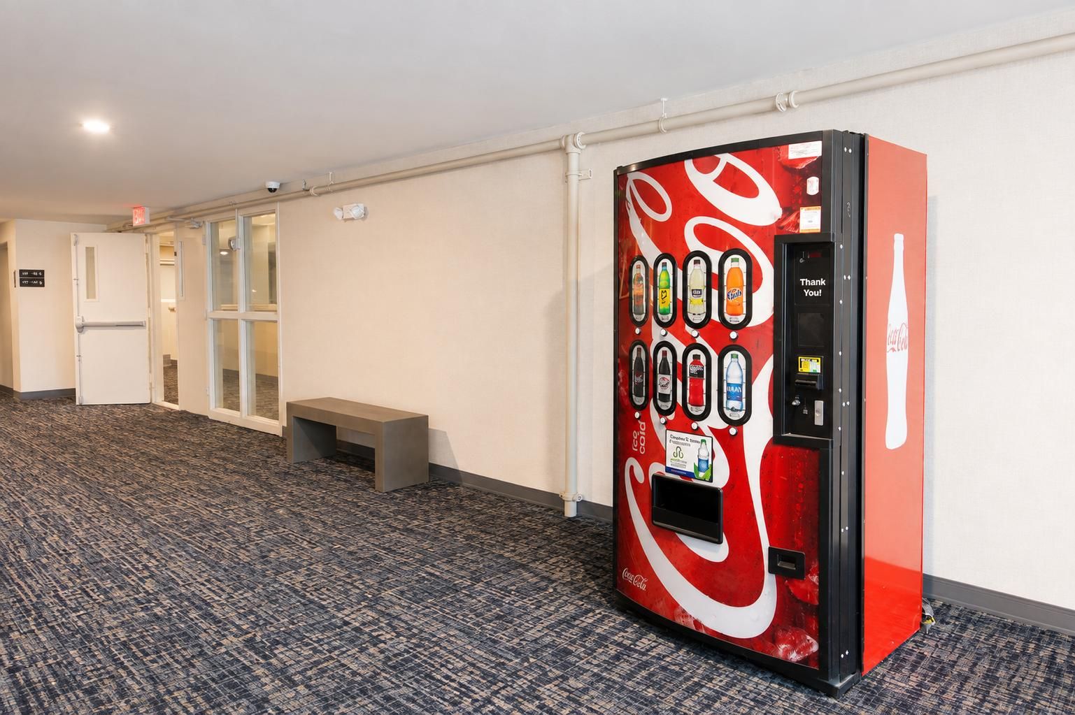 Un distributeur automatique proposant diverses boissons, un banc et une porte dans un couloir.