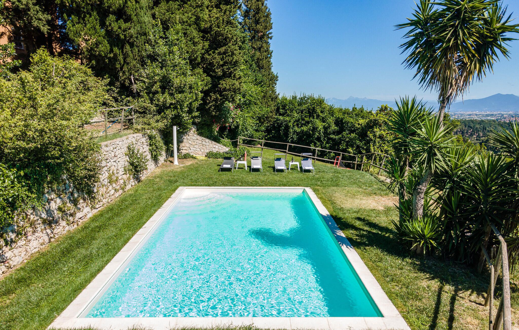 A rectangular swimming pool with clear blue water, surrounded by a grassy area and trees. There are five lounge chairs aligned along the pool's edge.