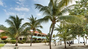 A beachside hotel with palm trees, a sandy courtyard, and a building with a red roof. - Beachfront 2BR Villa with Pool & Spacious Layout (Placencia)