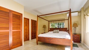 A four-poster bed with a wooden frame, a nightstand with a lamp, and a window with curtains. - Beachfront 2BR Villa with Pool & Spacious Layout (Placencia)