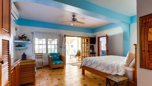 Un dormitorio con cama, una silla, una mesita, ventilador de techo y ventana.