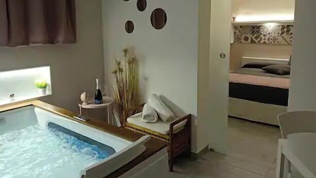 A room with a bathtub, a small table with a bottle, and a bed with a pillow. - Signorino Resort Double Room with Jacuzzi (MARSALA)