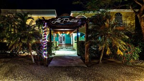 Una entrada con un letrero que dice "Los Porticos", rodeada de palmeras y luces decorativas.