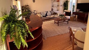 Ein Wohnzimmer mit einer Couch, einem Couchtisch und einem Fernseher. Pflanzen und ein Bücherregal sind ebenfalls vorhanden.