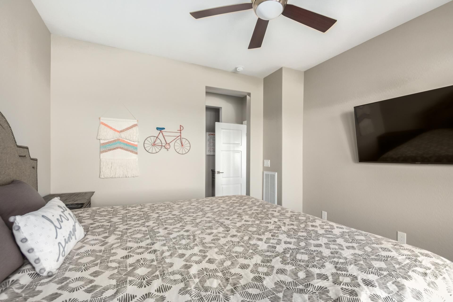 A bedroom with a bed, a ceiling fan, a wall-mounted TV, and a door leading to another room.