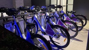 A row of electric bicycles with helmets and QR codes, likely for rental, in a modern indoor setting with a tiled floor and a brick wall.