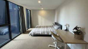 A modern hotel room with a large bed, a desk, and a view of the city through floor-to-ceiling windows.