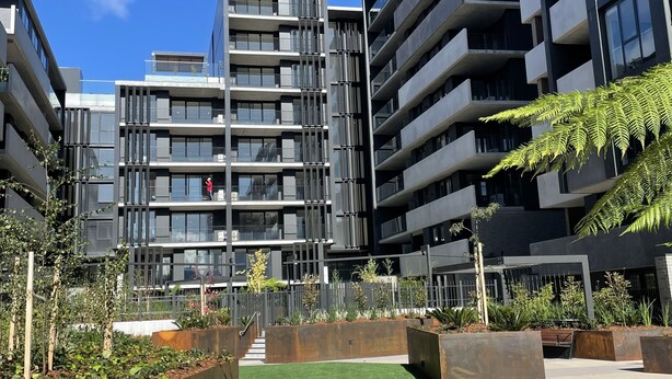 A modern multi-story building with balconies, a central staircase, and a landscaped area with artificial grass and planters.