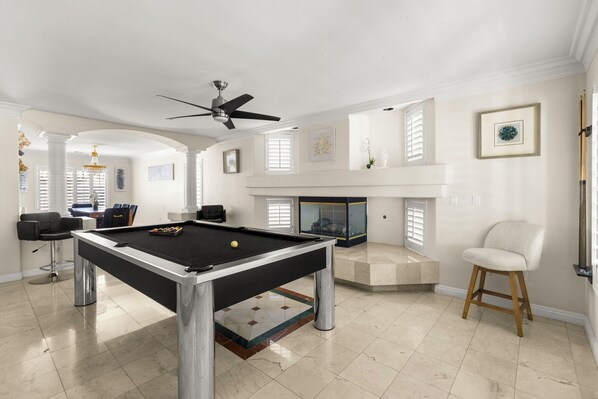 Une table de billard dans une pièce dotée d’un ventilateur de plafond, d’une cheminée et d’un fauteuil.