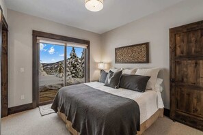 Un dormitorio con una cama grande, un ventanal corredero que da a una vista nevada del exterior, una puerta de madera y una obra de arte colgada en la pared.
