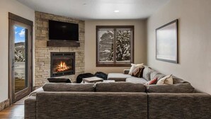 A living room with a stone fireplace, a large flat-screen TV, a sectional sofa, and a window with a snowy landscape view. - Ski-In/Ski-Out Updated Big Sky Retreat (Big Sky)