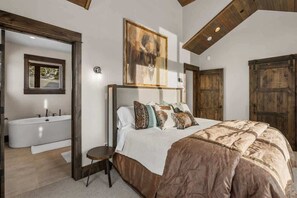 A bedroom with a large bed, a wooden headboard, a painting above the bed, and a view of a bathroom with a bathtub.