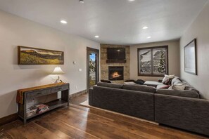 A modern living room with a fireplace, a large sectional sofa, a flat-screen TV mounted above the fireplace, and a painting on the wall. - Ski-In/Ski-Out Updated Big Sky Retreat (Big Sky)