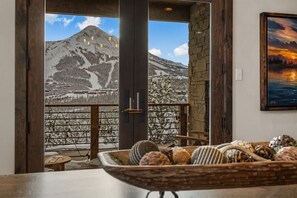 A room with a large window overlooking a snowy mountain landscape, a rustic wooden table with decorative items, and a framed painting on the wall.