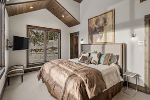 A bedroom with a large bed, a TV, a painting, and a window with a view of trees. - Ski-In/Ski-Out Updated Big Sky Retreat (Big Sky)