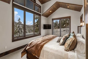 A bedroom with a large bed, two windows with mountain views, and a TV on the wall. - Ski-In/Ski-Out Updated Big Sky Retreat (Big Sky)