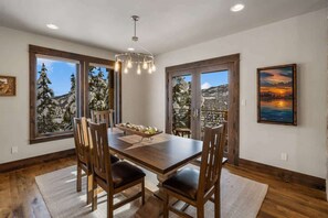 A dining room with a wooden table and chairs, large windows with a view of snow-covered trees, and a painting of a sunset. - Ski-In/Ski-Out Updated Big Sky Retreat (Big Sky)