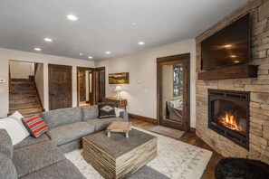 A modern living room with a stone fireplace, a large flat-screen TV, a gray sectional sofa, and a staircase leading to another level. - Ski-In/Ski-Out Updated Big Sky Retreat (Big Sky)