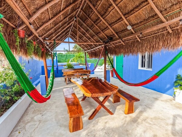 A covered outdoor seating area with wooden benches and tables, surrounded by blue walls and a thatched roof.