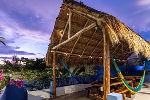A thatched-roof structure with a hammock and wooden furniture, surrounded by greenery and a view of the sky.