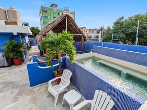 A rooftop pool area with blue tiled walls, a thatched-roof structure, and white lounge chairs.