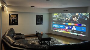 Uma sala de estar com uma televisão grande exibindo um filme, um sofá de couro, uma mesa de centro e um tapete estampado.