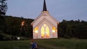 Une église dotée d’un clocher en flèche et de fenêtres en arc de cercle, entourée d’un champ verdoyant.