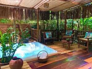 Una terraza de madera con un jacuzzi, sillas de madera con cojines estampados y abundante vegetación.