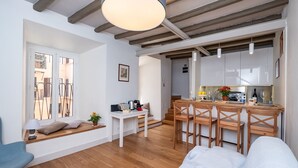 A modern apartment with a wooden floor, a dining area with a wooden table and chairs, a kitchen with white cabinets, and a large window with a view of buildings.