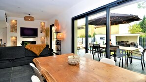 A modern living room with a large TV, a wooden dining table, and a glass sliding door leading to an outdoor patio area. - Stunning home in Montfavet (Montfavet)