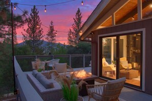 Una terraza al aire libre con mobiliario de mimbre, un brasero y luces colgantes con el atardecer de fondo.