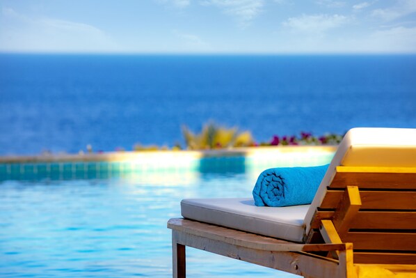 Un fauteuil de relaxation en bois, agrémenté d’une serviette bleue et d’un coussin blanc, est placé au bord d’une piscine offrant une vue sur l’océan.