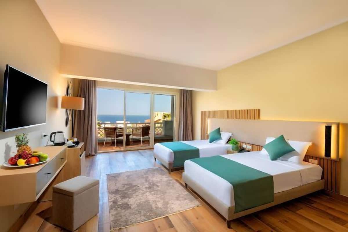Une chambre d’hôtel avec deux lits, une télévision, un bureau avec des fruits et une vue sur la mer.