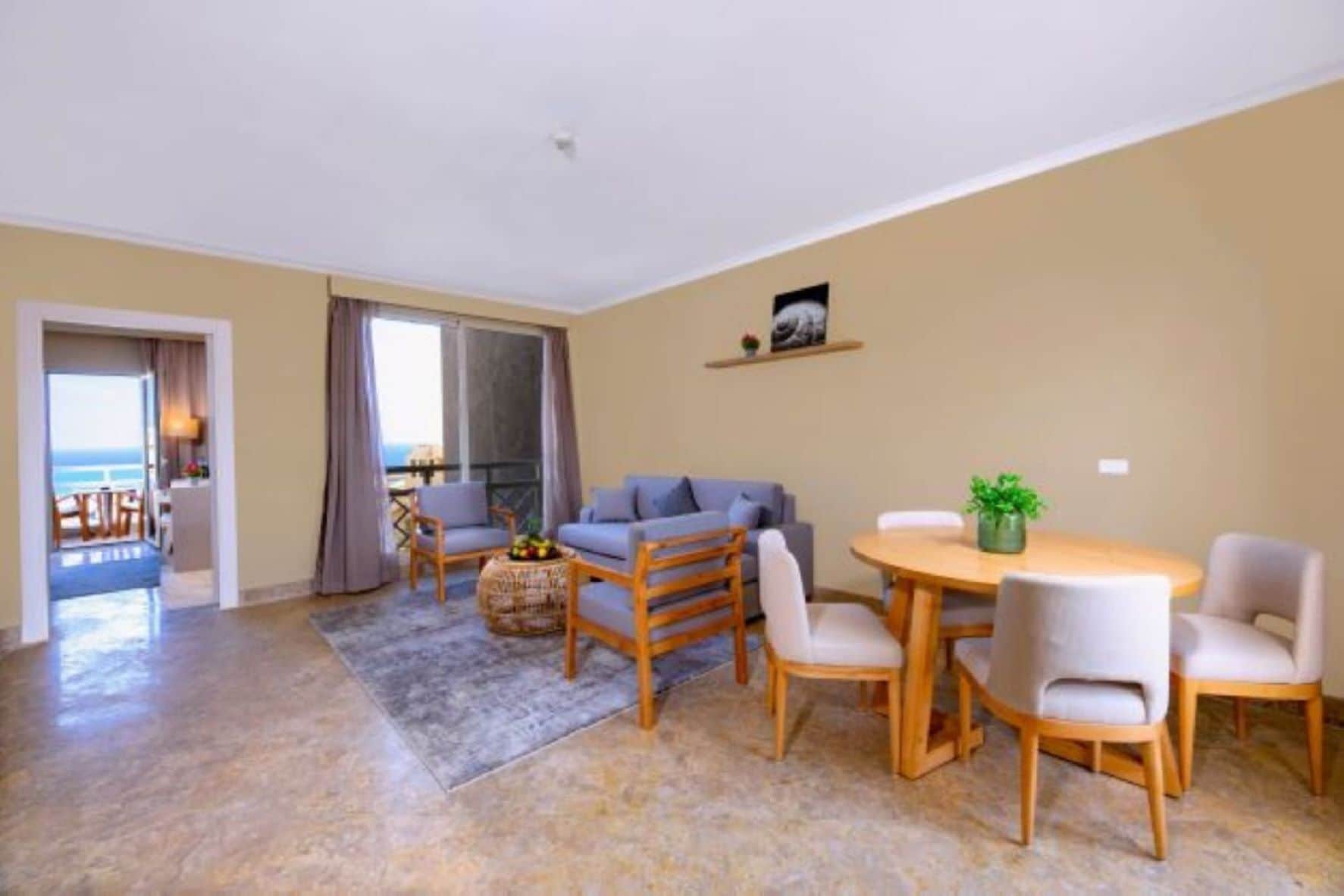 A spacious living room with a sofa, chairs, a coffee table, and a dining table. There is a balcony with a view of the sea.