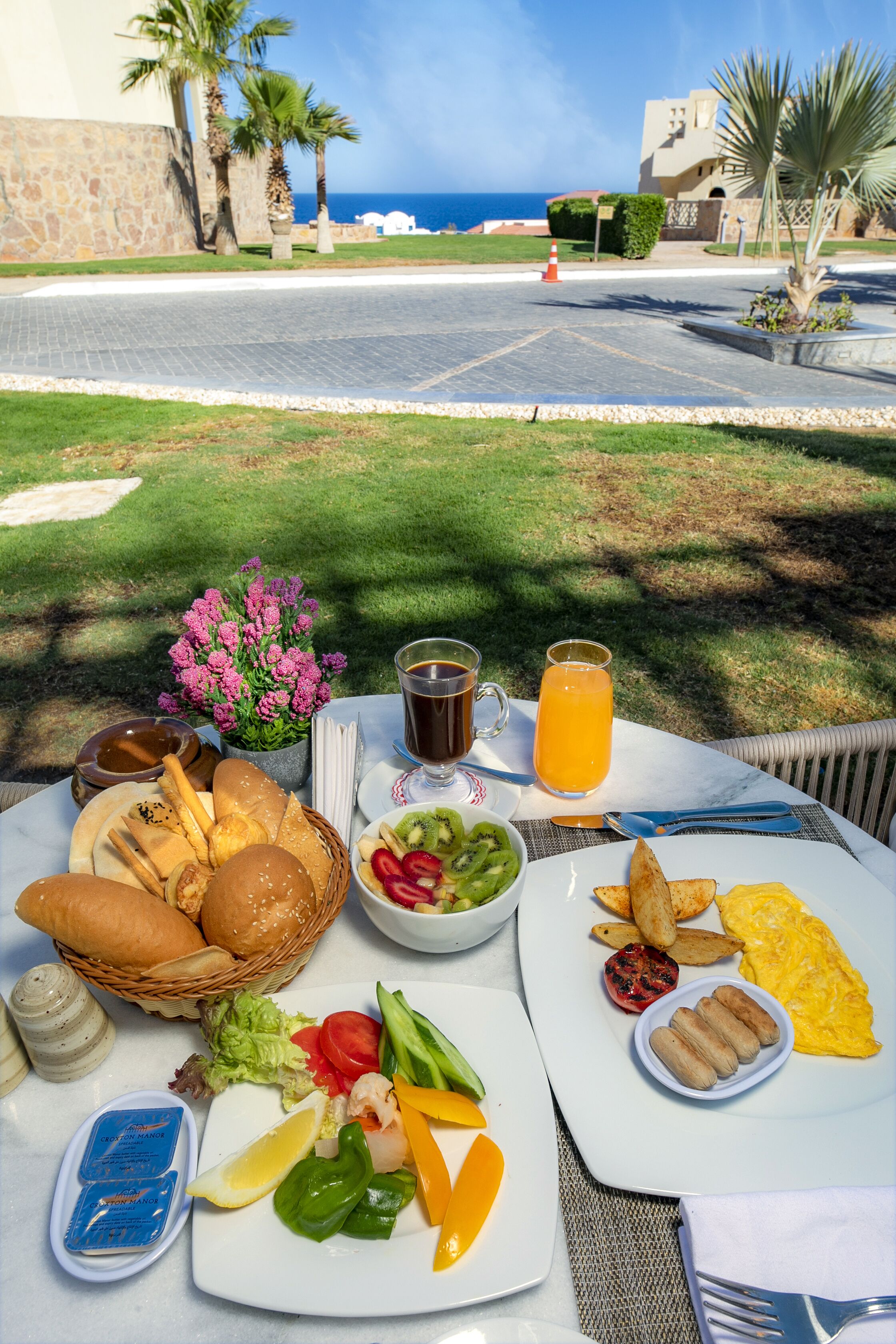 A table set with breakfast items including bread, eggs, and fruit, with a view of the ocean and palm trees in the background.