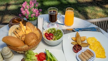 Una mesa dispuesta con alimentos para el desayuno, como pan, huevos y fruta, con vista al océano y palmeras al fondo.