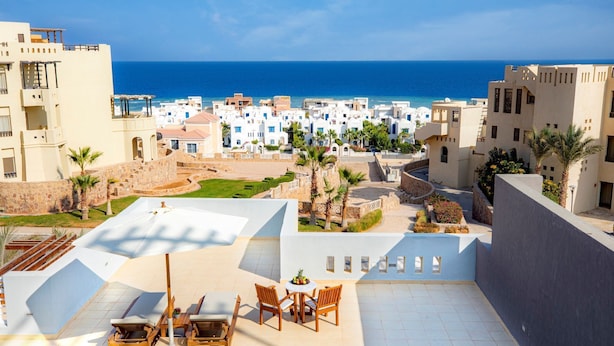 Une terrasse sur le toit offrant une vue sur la mer, aménagée avec des chaises et une table en bois, entourée de palmiers et d’un mur en pierre.